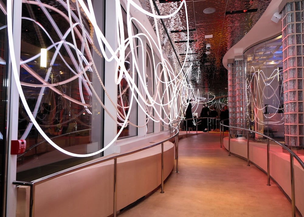 PERFECT TIMING, a site-specific light installation by Grimanesa Amorós on Wall Street, New York City, featuring suspended luminous sculptures that pulse and shift through warm golds, vibrant ambers, and subtle silvers, creating a rhythmic interplay of glowing light and shadow across the architectural space, evoking the flow of time and the energy of the urban environment in an immersive, radiant experience.