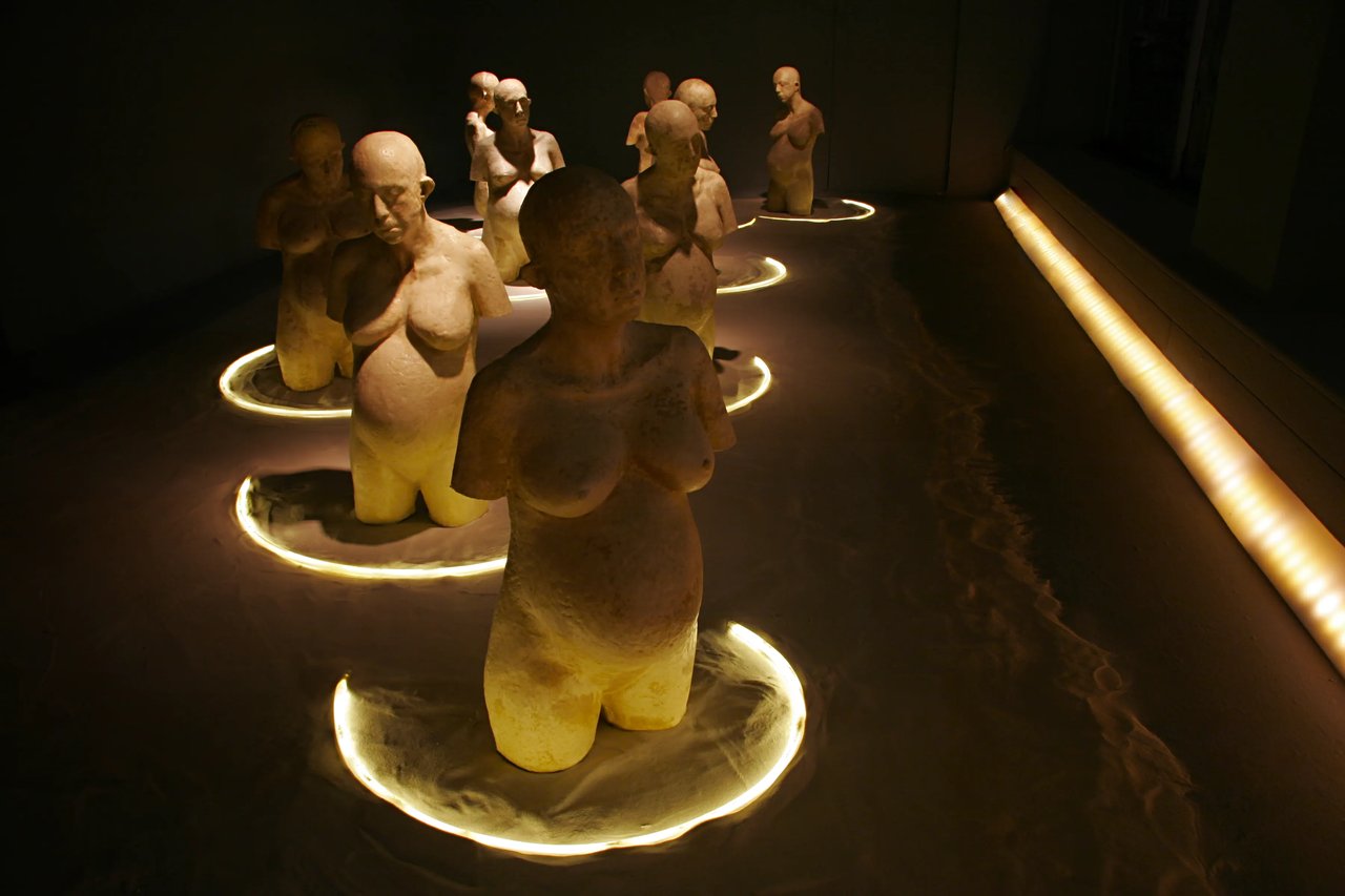 Grimanesa Amorós LA INCUBADORA, installation made of abaca cast paper and mixed media, each piece 39 x 19.5 x 13.5 inches, featuring handmade paper sculptures of pregnant female torsos with male heads cast from molds of the artist's own body, surrounded by soft pale sand, with lighting and music by composer Meshell Ndegeocello, exploring the relationship between biology, gender, and society. Exhibited at The Roger Smith Lab Gallery, New York, 2010.